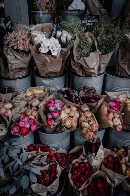 An array of fresh flower bouquets arranged in metal buckets and wrapped in brown craft paper, featuring a variety of roses in shades of pink, peach, and deep red, along with sprigs of eucalyptus and b