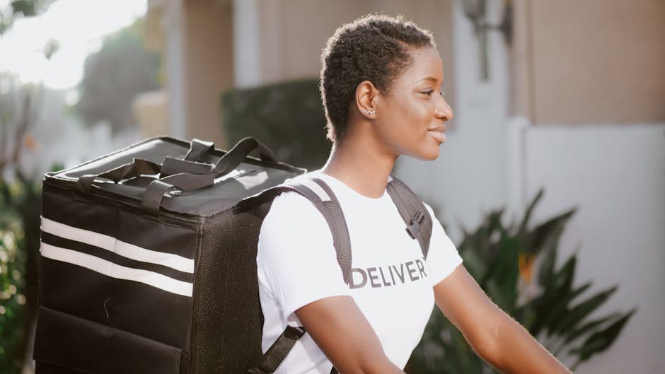A young woman with short curly hair, wearing a white t-shirt with 'DELIVERY' printed on it, is outdoors on a bright day. She is carrying a large black insulated delivery bag with white stripes on her 