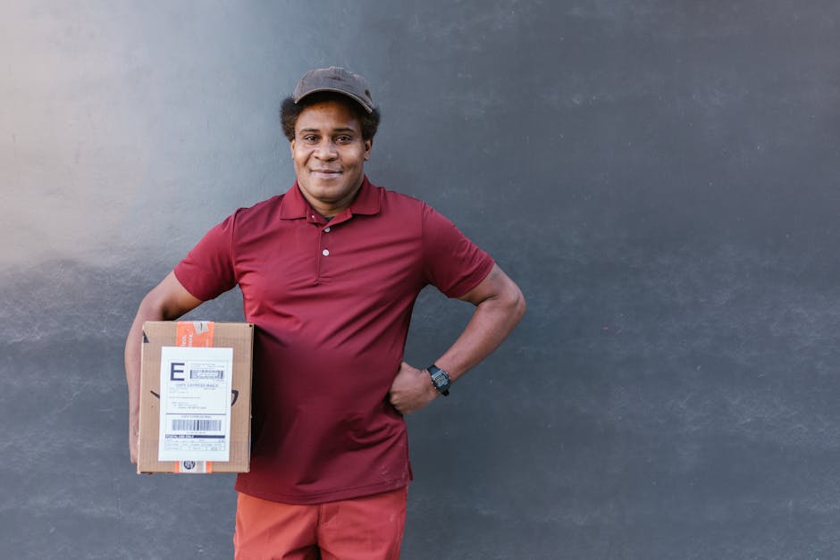 A male flower delivery courier with a friendly smile, wearing a burgundy polo shirt and matching cap, stands against a plain grey wall. He holds a small cardboard parcel with a shipping label in his r