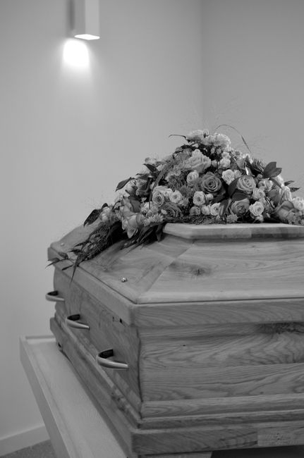 A wooden casket adorned with a floral arrangement featuring a mix of white and pale pink roses, lisianthus, and greenery, resting on a table in a funeral setting. The bouquet is placed on the top of t