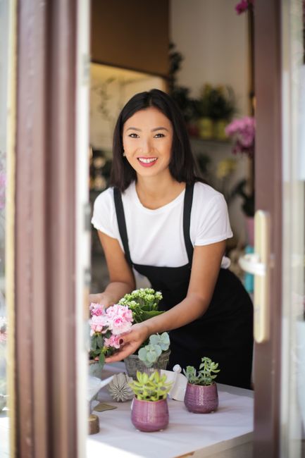 A young woman with shoulder-length dark hair and a bright smile is seen through a doorway, holding a fresh floral arrangement featuring pink carnations and white babies' breath. She is wearing a white