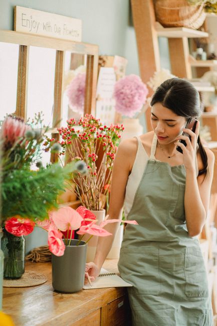 A young woman wearing a light green apron is standing in a flower shop, holding a mobile phone to her ear with a gentle smile. She is surrounded by vibrant floral arrangements, including pink anthuriu