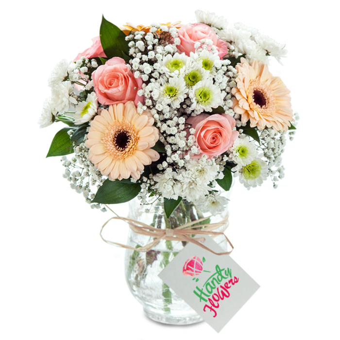 Elegant and softly radiant, this floral arrangement speaks to Norbury's blend of tradition and warmth. A cluster of luscious, pale peach-pink roses takes center stage, their velvety petals unfurling in tight spirals that speak of devotion and admiration. Siding the roses, two bold peach-orange gerbera daisies flash wide, textured petals and distinct dark centers-a playful nod to Norbury's colourful shops and parks. Between these, airy tufts of white baby's breath add delicate structure and fill, imbuing the bouquet with cloud-like volume. Rounding out the arrangement, petite white chrysanthemums with subtle lime-green hearts lend freshness and depth that mirrors the crisp green spaces surrounding Norbury's quiet residential roads. Deep green eucalyptus foliage is threaded throughout, its silvery-grey veins lending both texture and a gentle, clean scent. All rests within a classic, heavy-bottomed glass vase whose tapered neck and transparent form reveal healthy, upright stems immersed in water. Around the neck, a neat twine ribbon is tied, supporting a simple white tag for written well wishes. Photographed under softly diffuse daylight, the clarity and richness of each bloom are highlighted, making this bouquet a timeless Norbury gift-perfect for expressing gratitude, celebrating milestones, or simply sharing a piece of local natural beauty.