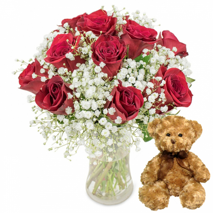 A bright, uncluttered studio-style photograph highlights a classic coupling of flowers and a keepsake teddy bear, thoughtfully arranged as if ready for same-day delivery across Norbury and nearby streets. On the left side, a clear glass vase holds a lush bouquet of approximately twelve deep crimson roses, each bloom rich with velvety petals. Some roses are fully open, their layered petals forming broad rosettes that reveal intricate spirals, while others remain half-open, adding a sense of unfolding romance. The colour is a saturated red with hints of burgundy in the shaded curves, giving the arrangement a dramatic yet refined character. Among the roses, delicate stems of white baby's breath weave softly through, each bearing tiny, cloud-like clusters of blooms. These flecks of white lighten the composition, creating a gentle, romantic contrast that recalls traditional Valentine's Day bouquets or anniversary flowers exchanged in a cosy Norbury home. Through the clear glass, the viewer can see the stems neatly gathered, their fresh green tones set against the still, transparent water. To the right of this elegant floral display, a plush teddy bear sits upright, its light brown fur looking wonderfully soft and touchable. The bear's simple features-small, round black eyes; a neat embroidered nose; and a faint, curved stitched smile-lend it an expression of quiet companionship. A dark brown satin-style ribbon is tied around its neck, catching a hint of light and echoing the deeper tones found in the roses. The bright white background and base create a calm, almost gallery-like setting where the rich reds, soft whites, and warm browns become the focal points. Together, this arrangement and teddy bear speak of affection, warmth, and thoughtfulness, making the scene perfectly suited to depict romantic surprises or heartfelt apologies delivered to loved ones living in and around Norbury.