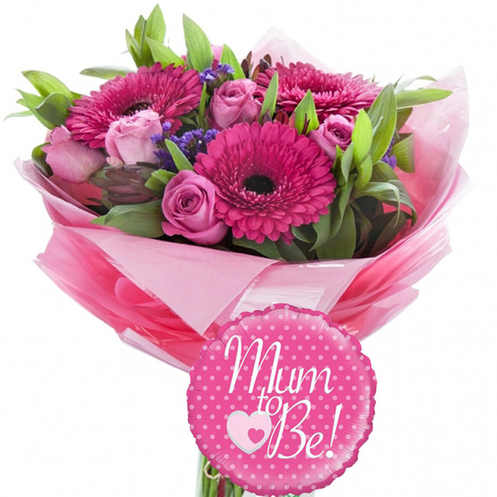 A celebratory "Mum to Be" flower arrangement is shown in the centre of the frame, set against a plain white background that enhances its lively pink palette, as if it has just been prepared at Florist Norbury for delivery to a local baby shower. The bouquet is composed in a classic rounded shape, dominated by vivid fuchsia gerbera daisies. Each gerbera features a rich ring of bright petals radiating from a deep, dark centre, giving the arrangement a bold visual anchor. Interspersed among them are multiple soft pink roses in different stages of bloom: some are small and tightly furled, suggesting promise and new beginnings, while others are partially open, revealing layers of satin-like petals that create a sense of depth and softness. Scattered between these main blooms are small clusters of purple filler flowers, possibly statice or limonium, adding a dusting of cool colour and a feathery texture that contrasts with the smooth petals of the gerberas and roses. Supporting the flowers is a bed of fresh green foliage, with leaves of varied shapes and rich tones, framing the blossoms and giving the bouquet a lush, full appearance. The stems are bound together and wrapped generously in multiple shades of pink tissue paper, flaring out in loose, ruffled folds around the base, then encased in a layer of clear, glossy cellophane that reflects light and emphasizes the bouquet's gift-like quality. In front of the flowers, a round fuchsia foil balloon with a gently scalloped edge catches the eye. Its surface is dotted with bright white polka dots and emblazoned with the phrase "Mum to Be!" in graceful white script, accompanied by a motif of a pink heart encircling a smaller white heart. The entire scene suggests joy, tenderness, and anticipation, the kind of thoughtful gift you might see carried along Norbury Avenue on the way to a family celebration.