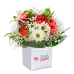 A lively floral arrangement beams with joy from within its crisp white gift bag, offering a taste of Norbury's welcoming vibrance. The bouquet weaves together exuberant coral, peach, and vivid red carnations, each bloom unfurling with velvety ruffles reminiscent of gardens found just off Green Lane. Dotted among them, snowy white daisies with their cheerful golden-green hearts sparkle like carefree laughter. Baby's breath graces the bouquet with its gentle, airy touch, while hints of purple-lavender buds add a subtle promise of things yet to come. Broad, glossy green leaves wrap around the blooms, grounding the arrangement in natural lushness. The bag's sturdy white ribbon handles and the pristine background create a sense of gift-giving anticipation, as if this display could travel from a Norbury florist straight to a loved one's doorstep. Ideal for any heartfelt moment-whether a birthday, new beginning, or simple thank you-this bouquet delivers a fresh, uplifting splash of Norbury's exuberant spirit right into the heart of the celebration.
